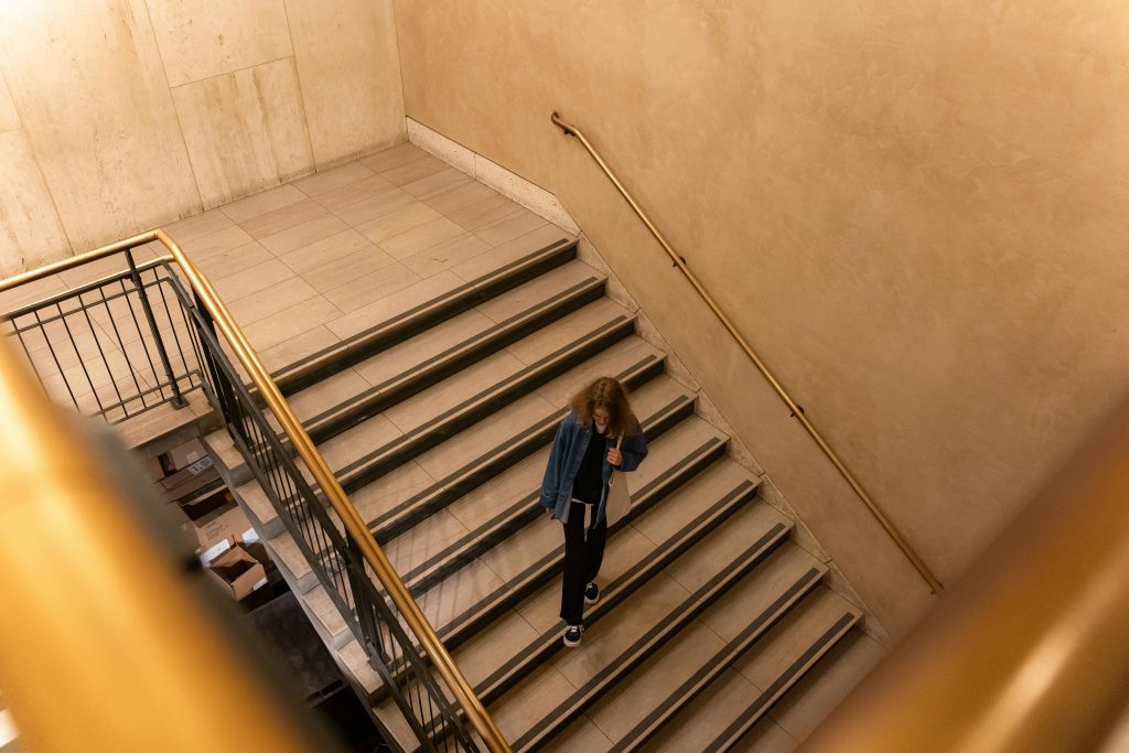 A person walks down a modern indoor staircase with beige walls and metallic railings.